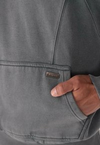 Main dans la poche avant d'un sweat-shirt gris avec une petite étiquette rectangulaire marron "Dropside" sur la poche.