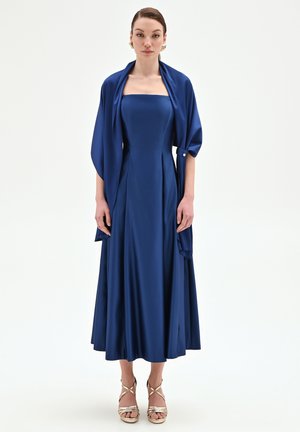 Donna con i capelli raccolti che indossa un abito midi blu navy senza spalline con uno scialle abbinato, completato da sandali con tacco alto dorati e sottili cinturini.