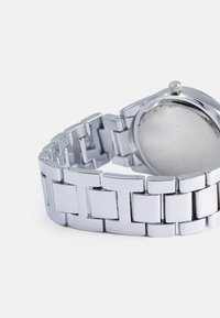 Ασημένιο ρολόι από ανοξείδωτο ατσάλι με γυαλιστερή φινιρίσματος, διαθέτοντας βραχιολάκι με ορθογώνιους συνδέσμους και στρογγυλή θήκη. Διακριτική σήμανση στο πίσω μέρος.