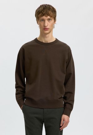 Jeune homme aux cheveux châtain clair portant un sweat-shirt marron foncé uni et un pantalon sombre, debout devant un fond clair uni.