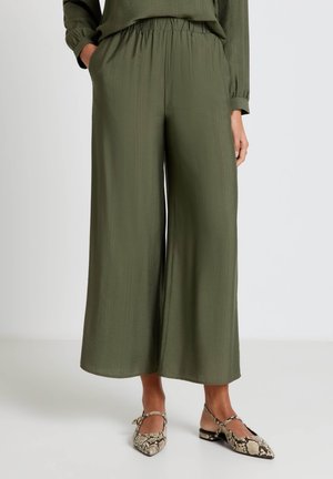 Donna che indossa pantaloni larghi a gamba corta di colore verde oliva con vita elastica e scarpe a punta con tacco basso e stampa pitone.