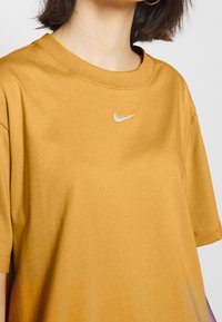 Bawełniany t-shirt w kolorze musztardowej żółci z okrągłym dekoltem, krótkimi rękawami i białym logo Nike umieszczonym na środku klatki piersiowej. Gładka tekstura, luźny krój.