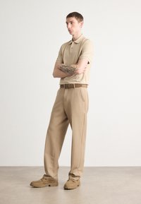 Khaki spalvos polo marškiniai ir plačiakelnės kelnės, papildytos derančiu diržu ir zomšos loafers, su lygia tekstūra ir neutralių spalvų palete.