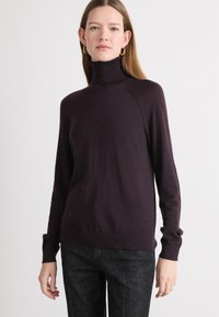 Un pull à col roulé bordeaux avec des manches longues, des poignets côtelés et une texture lisse, porté avec un jean à motifs sombres.
