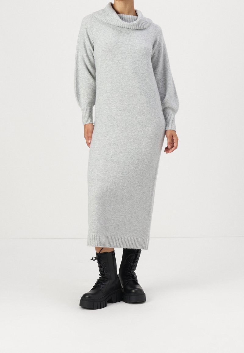 Personne portant une longue robe en maille gris clair avec un col bénitier et des manches bouffantes, associée à des bottes noires épaisses à lacets.