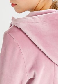 Närbild av en person som bär en mjuk rosa hoodie, som visar axeln, huvan och en del av ansiktet i profil.