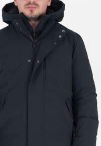 Donkerblauwe pufferjack met een hoge kraag en verstelbare capuchon, voorzien van ritsen en drukknoopjes. Getextureerd oppervlak met zijzakken.