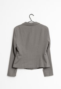 Harmaa kuviollinen bleiseri, jossa on lovettu kaulus ja pitkät hihat. Ominaisuuksiin kuuluu geometrinen muotoilu ja hieman vartalonmyötäinen siluetti. Roikkuu mustassa koukussa.