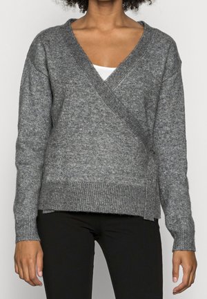 Personne portant un pull en maille gris style cache-cœur par-dessus un haut blanc et un pantalon noir, debout devant un fond clair uni.