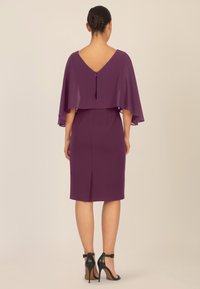 Violetti mekko, jossa on viittamainen päällyste ja V-mallinen selkäosa. Sileä kangas, polvipituinen leikkaus ja takaventti. Yhdistetty mustiin korkokenkiin.