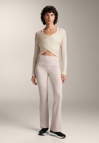 Hellbeige Wickelpullover mit gerippter Textur über einer rosa-hochgeschnittenen Leggings. Schwarze Sportschuhe vervollständigen das Outfit.