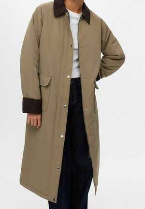 Persona con un abrigo largo beige con cuello y puños marrón oscuro, camisa blanca y vaqueros azul oscuro, de pie con una mano en la cadera.