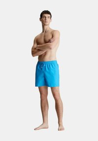 Costumi da bagno blu realizzati in tessuto leggero, con vita elastica e coulisse, e tasche laterali. Il modello sta in piedi a piedi nudi, con le braccia incrociate.