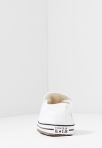 Baskets en toile blanches avec semelle en caoutchouc noir et bandes blanches ; présente le logo Converse sur le talon et un design à bout rond. Tissu supérieur texturé.