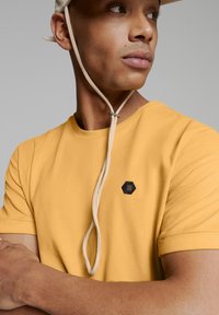 Geel katoenen t-shirt met korte mouwen, voorzien van een zwart hexagonaal logo. Het model draagt een crèmekleurige muts met verstelbare banden.