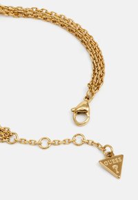 Goldene Metallkettenschnur mit ineinander verwobenen Ketten, einem Karabinerhaken und einem dreieckigen Anhänger, der mit "GUESS" graviert ist.
