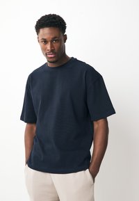 Young man with short curly hair wearing a dark navy t-shirt and light beige pants stands with hands in pockets against a plain white background.