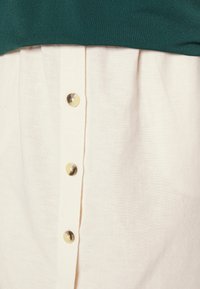 Blusa abotoada com a parte superior verde e a parte inferior creme. Apresenta cinco botões creme, textura plissada e um design em camadas.