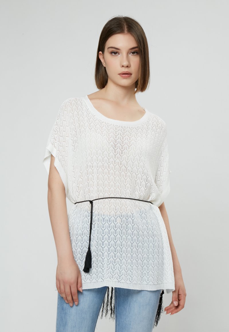 Witte, losvallende top met golvend textuurpatroon, korte mouwen en een zwarte trekkoordriem met kwastjes; gecombineerd met blauwe jeans.