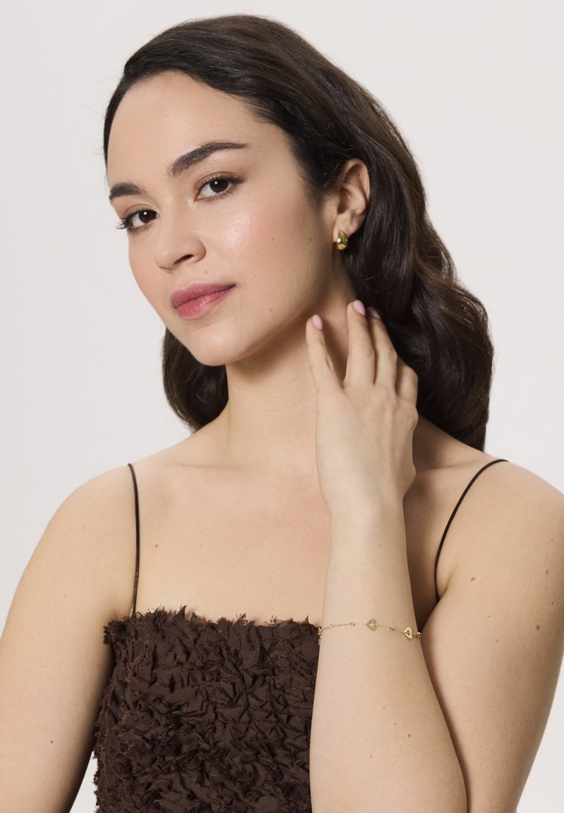 Złote kolczyki w kształcie kół, brązowa strukturalna bluzka oraz delikatna złota bransoletka z małymi charmsami. Modelka pozuje z jedną ręką na szyi.