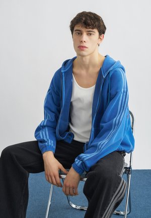 Jeune homme portant un sweat à capuche bleu zippé et un débardeur blanc, assis sur une chaise en métal devant un fond uni.