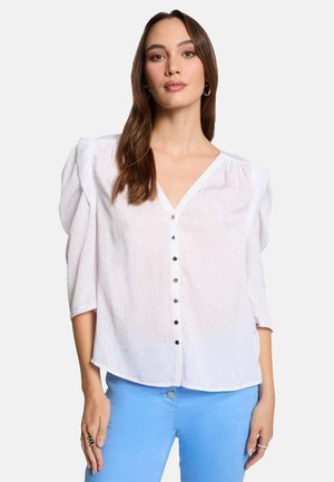Blusa bianca, strutturata, con scollatura a V, maniche corte a palloncino e chiusura con bottoni. Abbinata a pantaloni in denim blu.