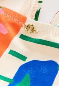 Gros plan sur un tissu coloré avec des formes orange, vertes et bleues, une bordure en mesh crème, une couture en zigzag et une petite boucle métallique circulaire dorée.