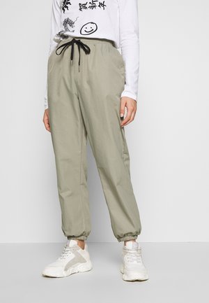 Pantalon de jogging beige décontracté avec cordon noir, poignets élastiques, porté avec des baskets blanches et un haut blanc à manches longues avec motifs noirs.