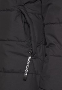 Chaqueta acolchada negra con textura de acolchado, que presenta un bolsillo con cremallera y una etiqueta de tela que dice "DIDRIKSONS". Material suave.