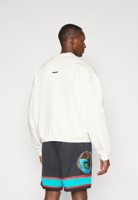 Hvid sweatshirt med ribbet kant og manchetter, har et lille logo-tag på ryggen; parret med sorte shorts, prydet med et bjørne-graphic og farvede striber.