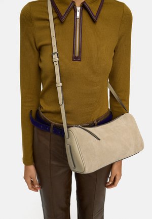 Kobieta ubrana w musztardowy prążkowany top z długim rękawem, suwakiem i skórzanym kołnierzem, brązowe spodnie, niebieski pasek oraz beżowa zamszowa torba na ramię.