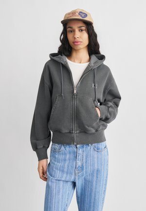 Žena v béžovej šiltovke „Heart of Detroit“, sivá mikina na zips s vreckami vpredu, biele tričko a svetlomodré pruhované džínsy, stojaca na bielom pozadí.