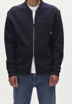 Homme portant un cardigan bleu marine foncé zippé avec deux poches basses et une poche poitrine, sur une chemise blanche et un jean bleu.
