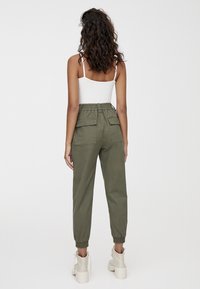 Pantalon cargo à taille élastique vert olive avec poches latérales et bas effilés. Porté avec un débardeur blanc et des bottines à plateforme de couleur claire.