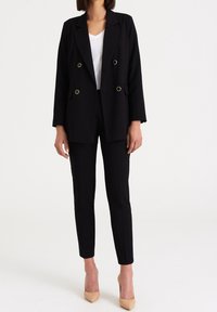 Blazer noir croisé avec des boutons dorés, associé à un pantalon noir taille ajustée et des talons beiges, sur un fond uni.