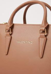 Beige keinokas laukku, jossa on kaksi yläpitoa, kultaiset yksityiskohdat, teksturoitu pinta ja kohokuvioitu "VALENTINO" -logo etupuolella.
