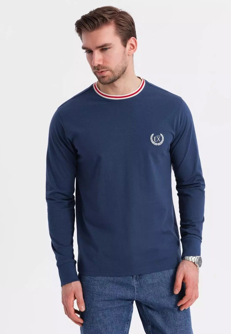 Camisa de manga larga azul marino con cuello redondo, que presenta detalles a rayas rojas y blancas. Incluye un pequeño logo bordado en el pecho.