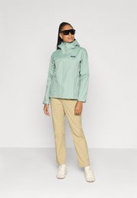 Lys mintgrønn vanntett jakke med hette, kombinert med beige bukser og hvite sneakers. Har glidelås foran og et logopatch.