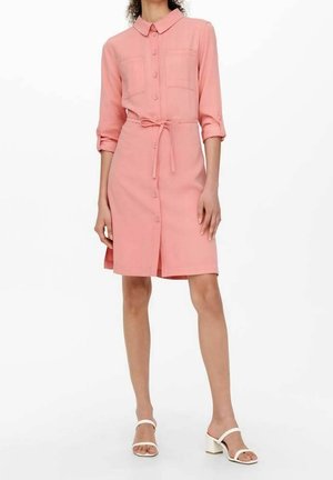 Robe boutonnée en tissu rose clair avec col, deux poches poitrine et ceinture à nouer. Texture lisse, longueur au genou, manches roulées, associée à des sandales blanches.