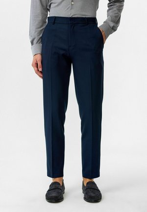 Marineblauwe broek met een getailleerde pasvorm, met een soepele textuur, flat front en zijzakken. Draagt met een grijs overhemd met lange mouwen.