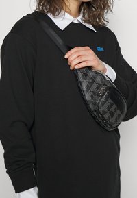 Bolso bandolera negro hecho de material texturizado, con un patrón geométrico, cierre de cremallera y un pequeño bolsillo delantero con un diseño elegante.