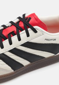 Białe sportowe buty skórzane z czarnymi paskami i czerwonymi akcentami. Wyposażone w czarne sznurówki, teksturowaną podeszwę oraz logo "PREDATOR".