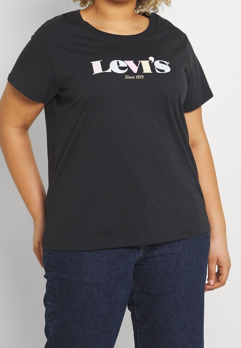Persoon in een zwart Levi's t-shirt met multicolor logo en blauwe jeans, staande met één hand achter hun rug.
