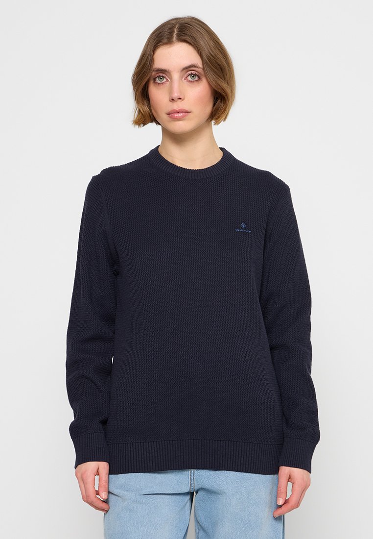 Granatowy sweter dzianinowy z okrągłym dekoltem, ściągaczowymi mankietami i dołem, z małym haftowanym logo na piersi oraz teksturowanym materiałem.