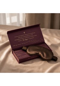 Maschera per gli occhi marrone in seta che riposa su una scatola viola aperta con testo dorato, posizionata su tessuto beige di raso sotto una luce naturale soffusa.