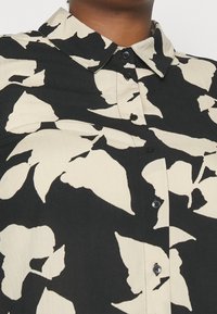 Camicia a bottoni con uno sfondo nero e grandi motivi di foglie beige. Il tessuto è leggero, con una texture liscia e colletto.