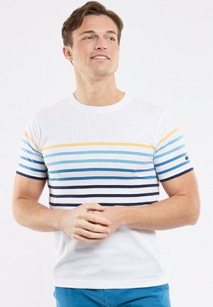 Homme portant une chemise blanche à manches courtes avec des rayures horizontales bleues et jaunes, les mains jointes, regardant légèrement vers le haut et sur le côté.