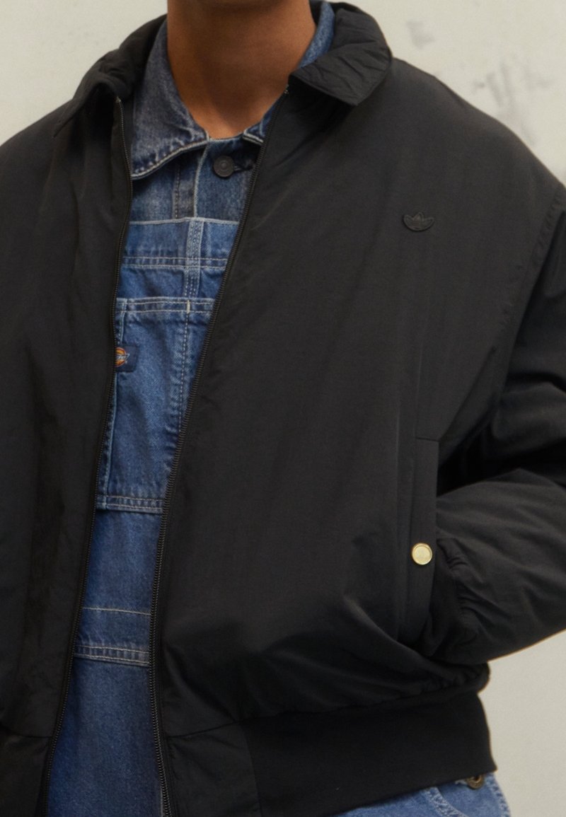 Černá lehká zipová bunda s kapucí, která má jemné logo, elastické manžety a hladký povrch přes džínsové overall.