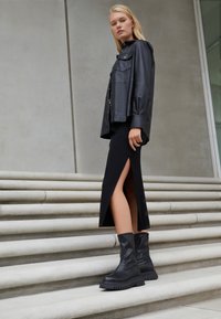 Donna bionda in piedi su scalini di pietra, indossando una giacca di pelle nera, una gonna midi nera con spacco laterale e stivali chunky neri alla caviglia.