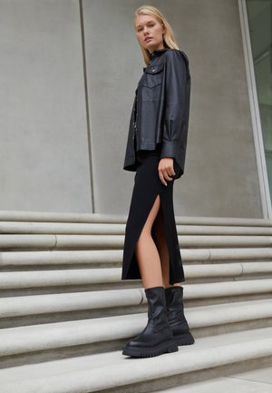Donna bionda in piedi su scalini di pietra, indossando una giacca di pelle nera, una gonna midi nera con spacco laterale e stivali chunky neri alla caviglia.